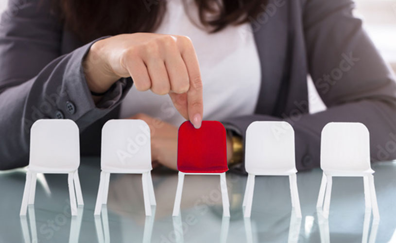 Geschäftsfrau wählt mit den Fingern einen roten Miniaturstuhl aus einer Reihe von weißen Miniaturstühlen.